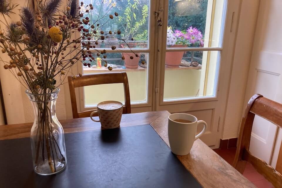 Zwei Kaffeetassen stehen auf altem Holztisch mit einem Trockenblumenstrauss. Im Hintergrund Fenster eines alten Hauses.
