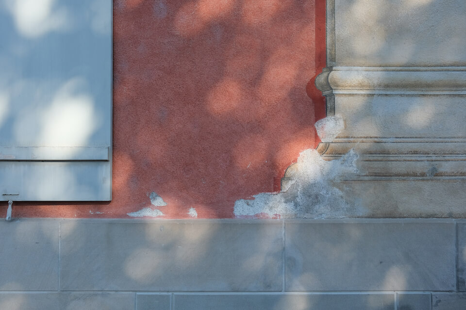 Nahaufnahme einer Fassade mit grauem Steinsockel. Der Putz darüber ist himbeerrot und mit einer Malerei eines Pilasters verziert. Auf der Fassade sind die Sonnen- und Schattenflecken eines Baumes sichtbar.